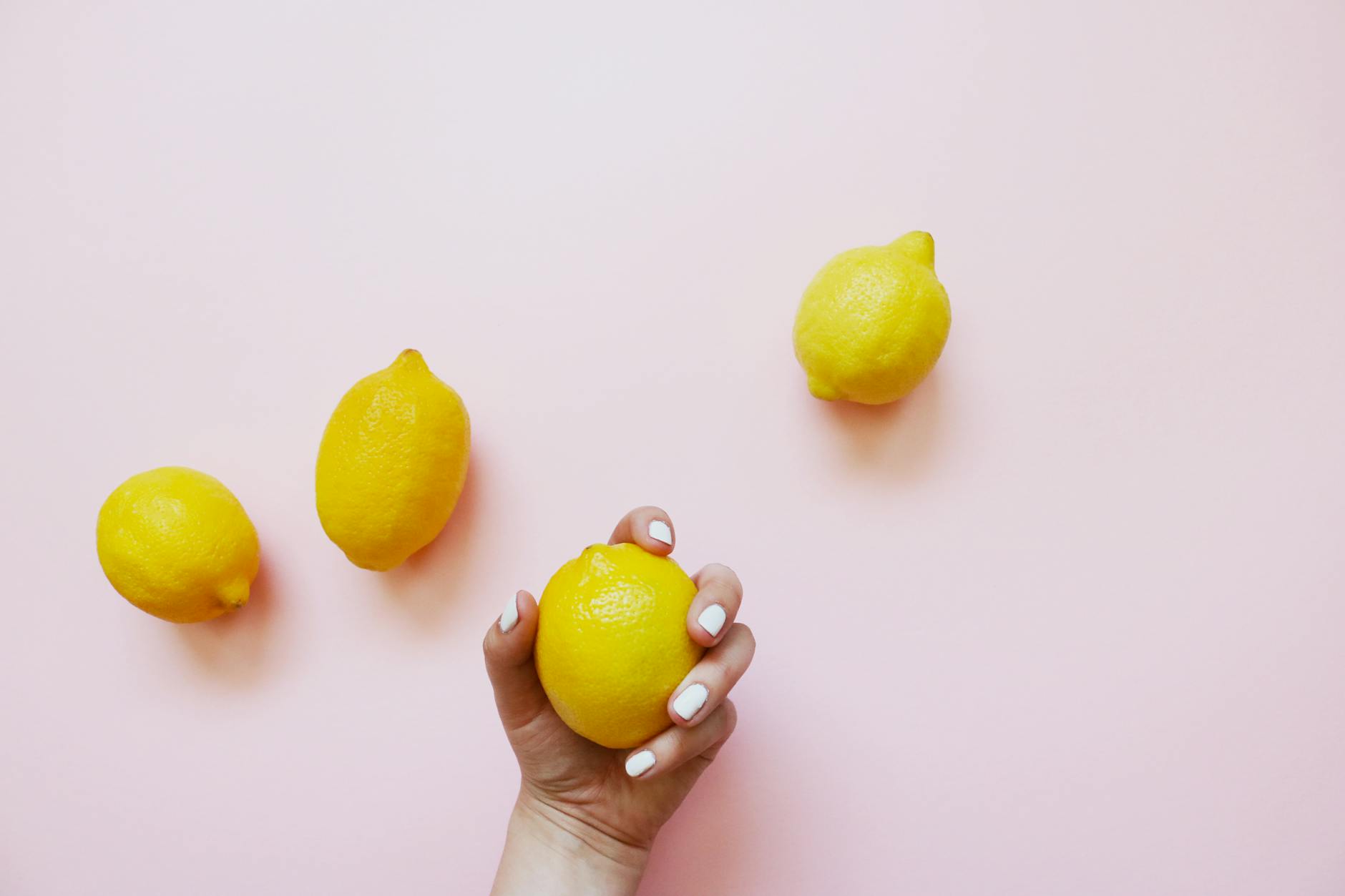 Mano con unghie bianche che tiene un limone su sfondo rosa morbido, circondata da altri limoni