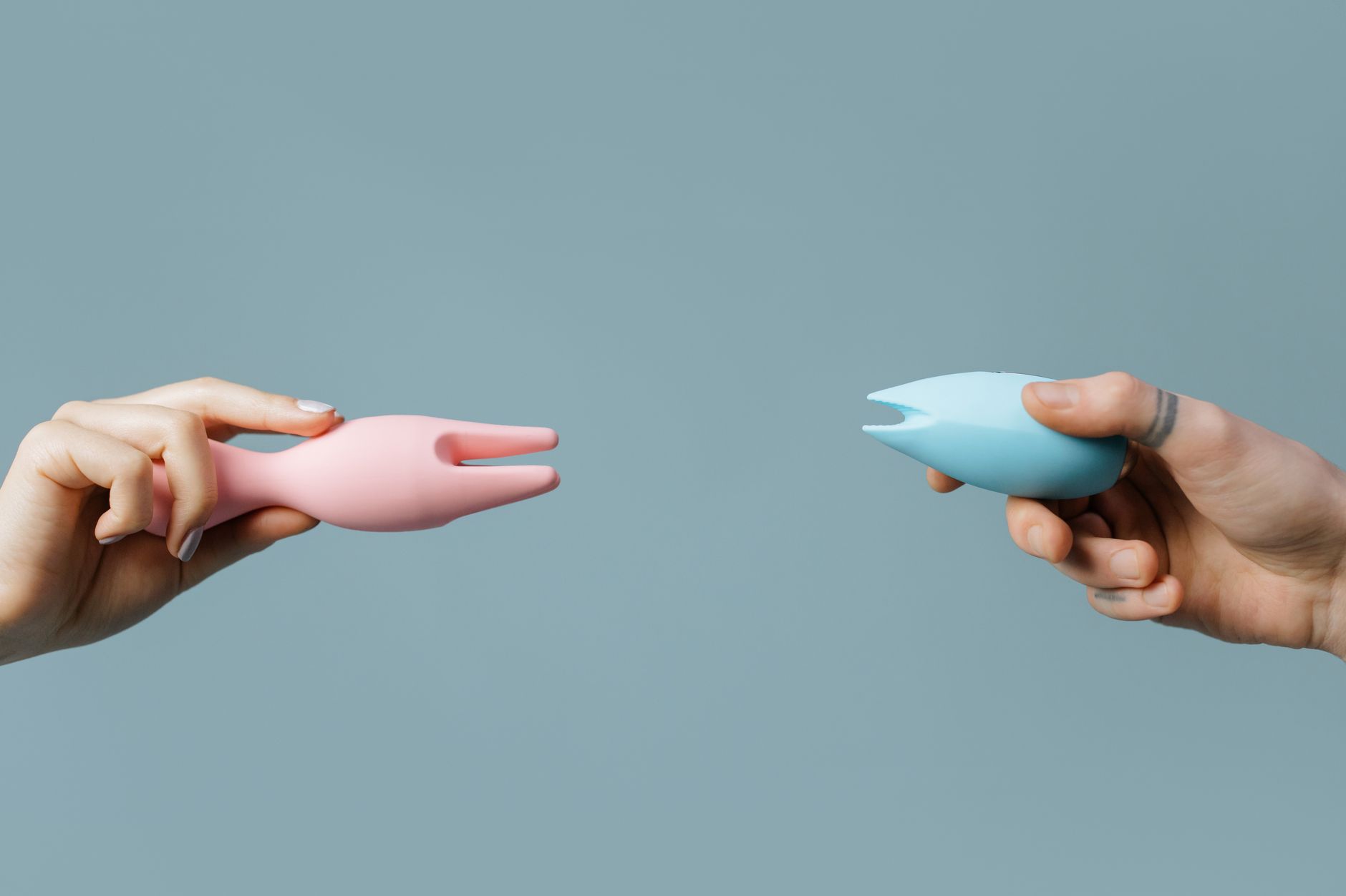Due mani che tengono vibratori in silicone rosa e blu su uno sfondo pastello soft