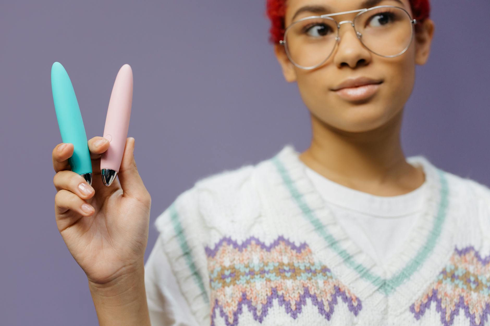 Una donna con occhiali colora tiene due vibratori colorati contro uno sfondo viola