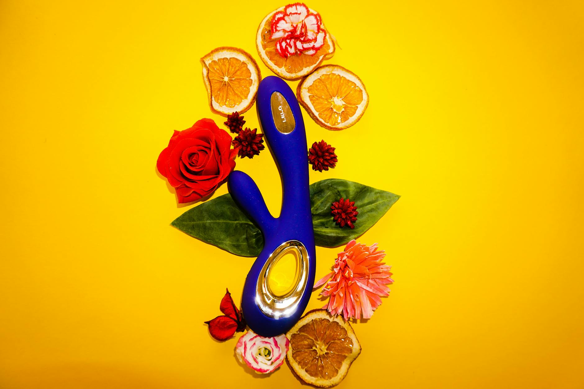 Vibratore blu circondato da fiori e fette d'arancia su sfondo giallo