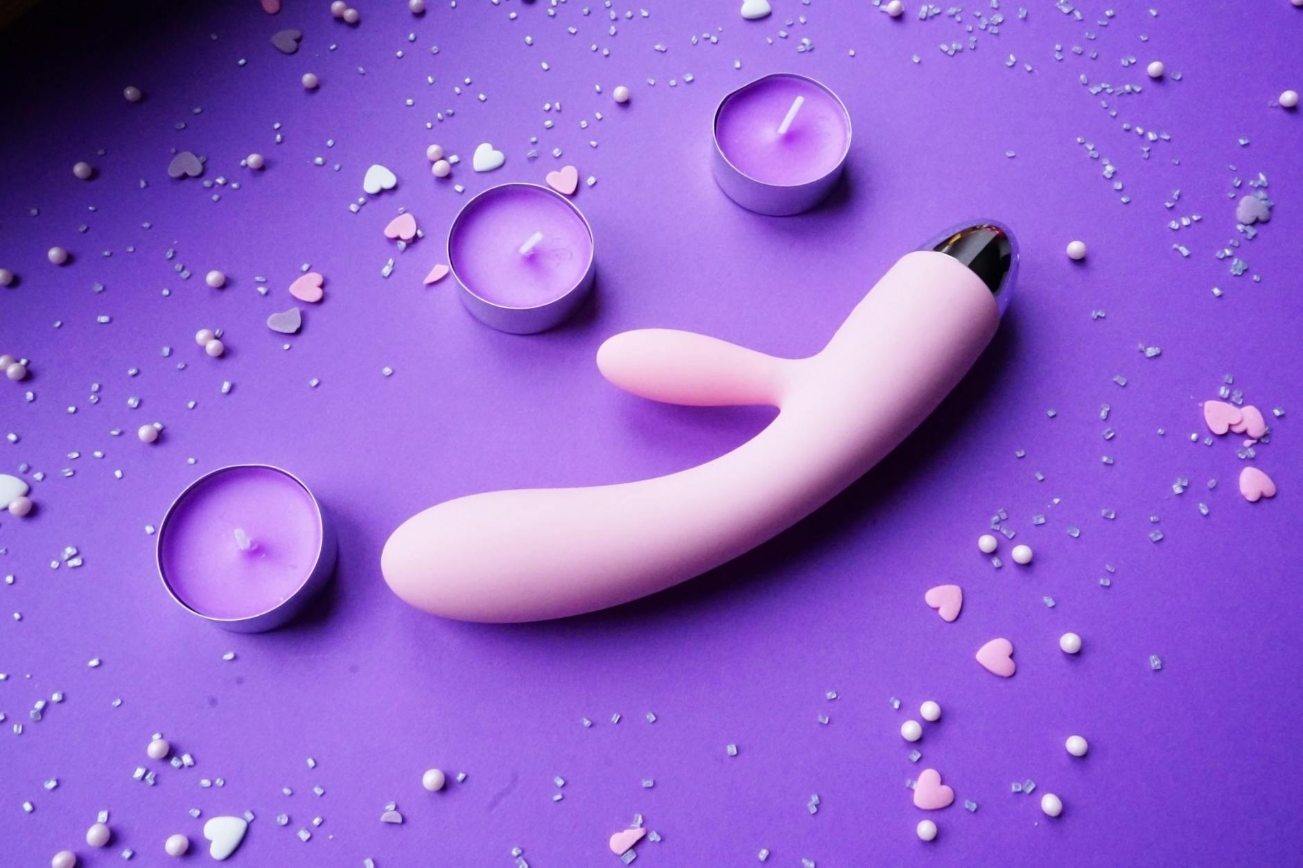 Vibratore rosa su sfondo viola con coriandoli a forma di cuore e candele per un'atmosfera romantica a distanza